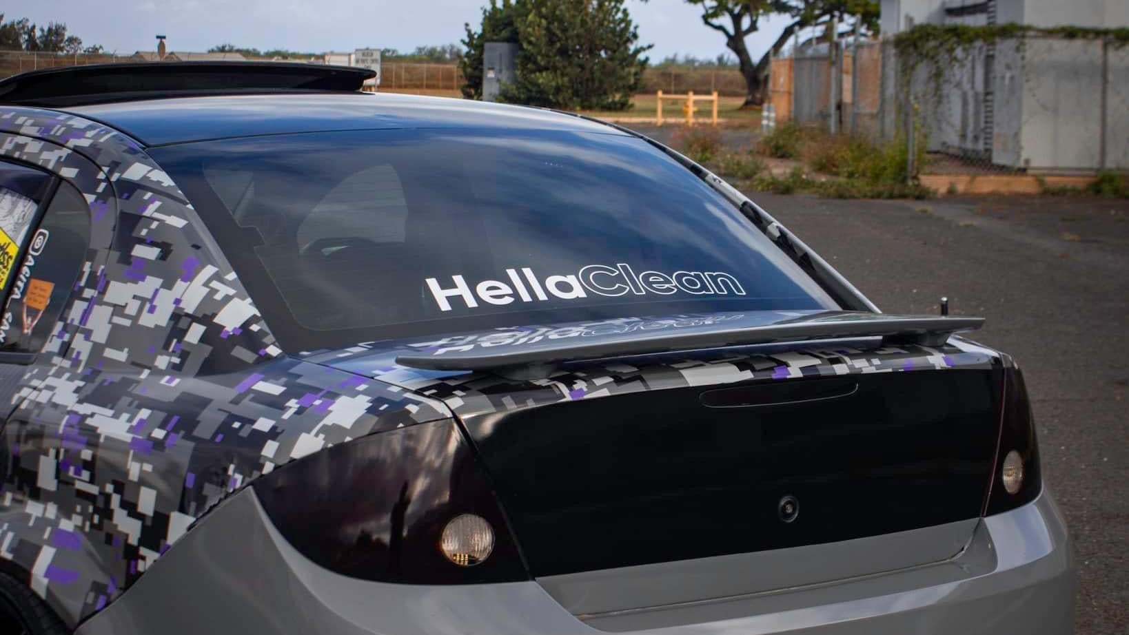 Car with 'Hella Clean' decal on a road with trees and buildings in the background